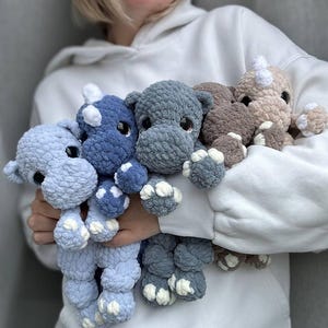 Puede incluir: Cuatro juguetes de peluche de rinoceronte tejidos a crochet en diferentes tonos de azul y marrón. Los juguetes están en los brazos de una persona que lleva una sudadera con capucha blanca.