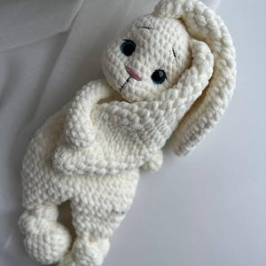 May include: White crocheted bunny plush toy with blue eyes and a pink nose. The bunny is curled up in a sleeping position.