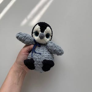 May include: A crocheted penguin plush toy featuring a soft grey body, black head markings, and white face accents. The toy has large blue plastic eyes, a small grey beak, and black feet. A thin dark blue ribbon is tied around its neck.