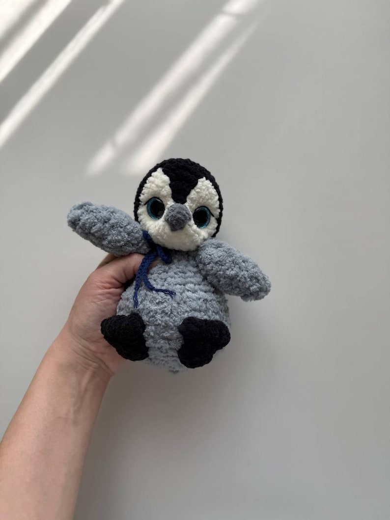 May include: A crocheted penguin plush toy featuring a soft grey body, black head markings, and white face accents. The toy has large blue plastic eyes, a small grey beak, and black feet. A thin dark blue ribbon is tied around its neck.
