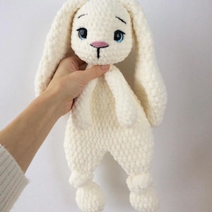 Peut inclure: Une poupée lapin en crochet blanc cassé faite à la main, avec de longues oreilles tombantes et une expression douce. Le lapin a des yeux bleus, un nez rose et est tenu par une personne. La peluche est douce et câline, idéale pour une chambre d'enfant ou comme cadeau.
