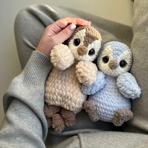 May include: Two plush penguin toys made of soft, textured yarn. One penguin is beige and brown with large dark eyes, while the other is light blue and white with similar dark eyes. Both have small wings, round bodies, and brown feet.