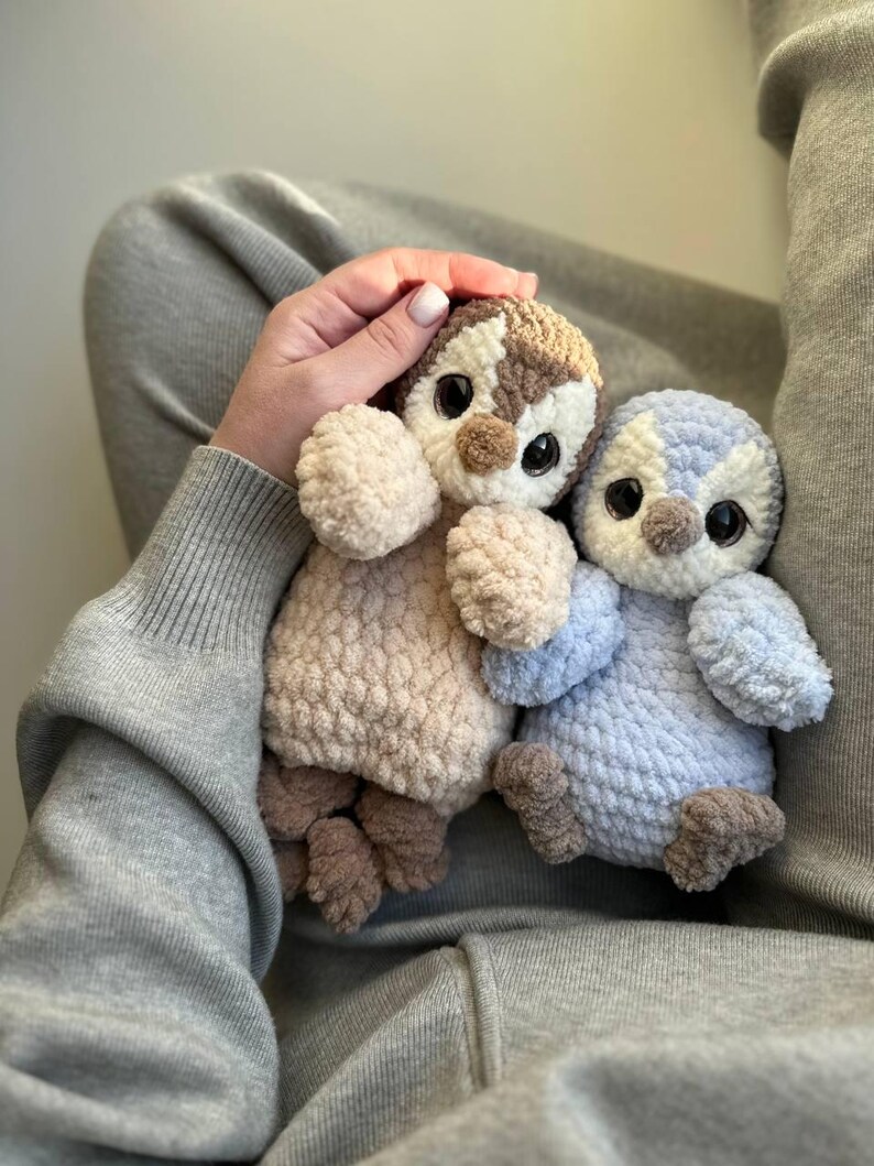May include: Two plush penguin toys made of soft, textured yarn. One penguin is beige and brown with large dark eyes, while the other is light blue and white with similar dark eyes. Both have small wings, round bodies, and brown feet.