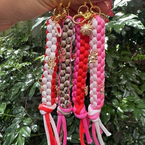 May include: Four braided keychains in red, white, pink, and brown with gold-coloured hardware. Each keychain features decorative charms, including hearts, crowns, and butterflies. The keychains have knotted tassels at the bottom.
