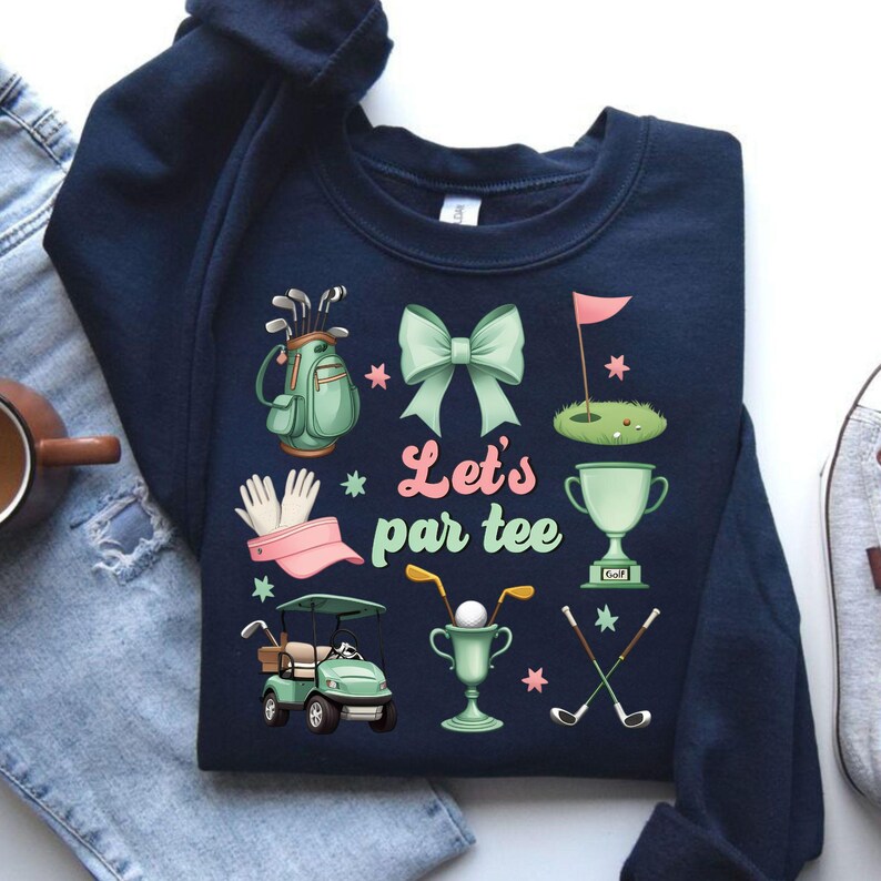 May include: Navy blue sweatshirt with a golf-themed design. It features a golf bag, golf clubs, a golf cart, a trophy, a golf ball, and a bow. The text "Let's par tee" is printed in pink on the sweatshirt.