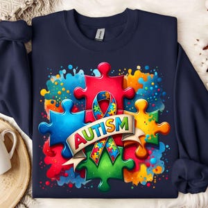 May include: A navy blue sweatshirt with a colorful design featuring puzzle pieces, a ribbon, and the word "AUTISM". The design is surrounded by watercolor-like splashes of color.