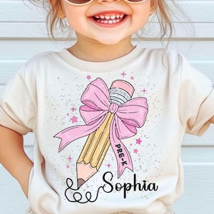 May include: Cream-colored t-shirt featuring a large pencil design with a pink bow and the text "PRE-K". The pencil is yellow with a pink eraser. The name "Sophia" is written below the pencil in a cursive font. Pink stars are scattered around the design.