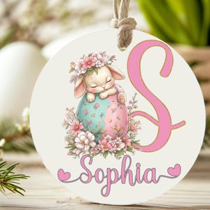 Könnte beinhalten: Runder cremefarbener Osterschmuck mit einem schlafenden Hasen mit Blumenkranz, eingebettet in ein pastellfarbenes Ei. Ein großer rosa Buchstabe "S" und der Name "Sophia" sind ebenfalls abgebildet. Der Schmuck hat eine natürliche Kordelaufhängung.