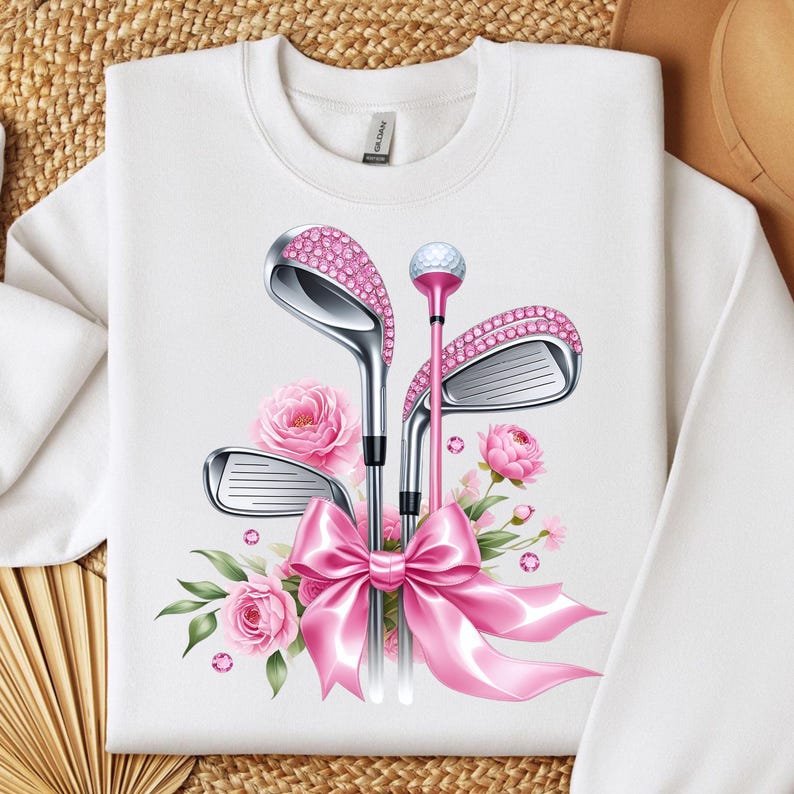 May include: White sweatshirt with a pink and silver design of golf clubs, a golf ball, and flowers. The golf clubs are decorated with rhinestones and tied together with a pink ribbon.