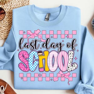 Puede incluir: Sudadera azul claro con el texto "&uacute;ltimo d&iacute;a de escuela" en letras rosas, amarillas, azules y negras. Las letras est&aacute;n decoradas con patrones de cuadros, rayas y lunares. El texto est&aacute; rodeado por un borde de cuadros rosa y blanco.