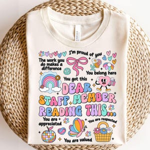 May include: A white t-shirt with a colorful graphic design that says "Dear Staff Member Reading This... You are appreciated. You are respected. You are valued." The graphic includes a rainbow, a butterfly, a peace sign, a heart, and flowers.
