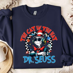 Peut inclure: Un sweat-shirt bleu marine avec un motif repr&eacute;sentant le Chat dans le Chapeau portant un casque et un n&oelig;ud papillon rouge. Le texte "The Cat in the Hat" est inscrit dans un cercle blanc avec une bordure &agrave; carreaux noirs et blancs. Le texte "Dr. Seuss" est en bleu sous le cercle.