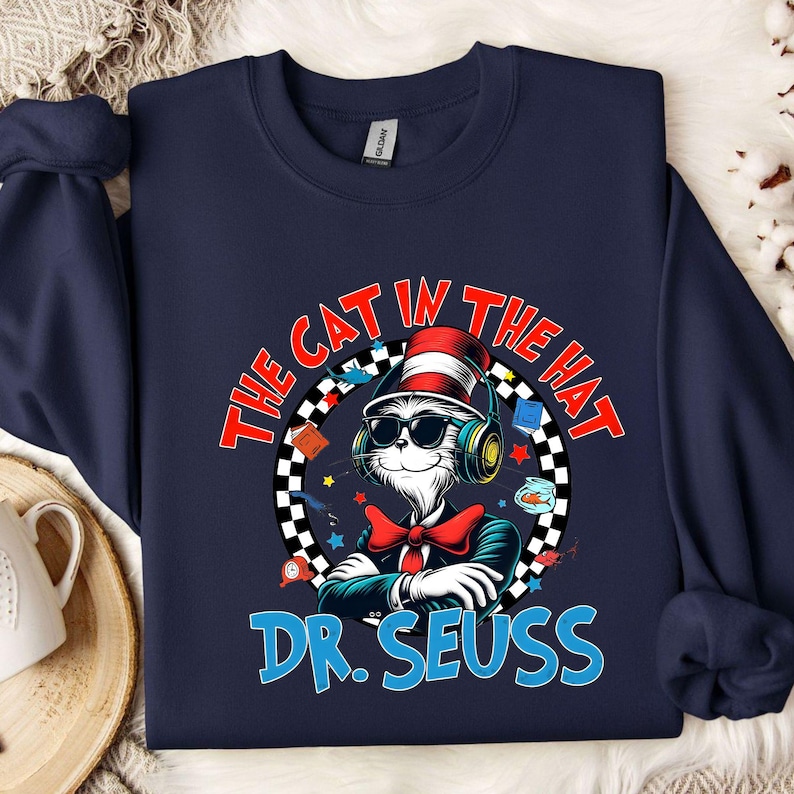 Peut inclure: Un sweat-shirt bleu marine avec un motif repr&eacute;sentant le Chat dans le Chapeau portant un casque et un n&oelig;ud papillon rouge. Le texte "The Cat in the Hat" est inscrit dans un cercle blanc avec une bordure &agrave; carreaux noirs et blancs. Le texte "Dr. Seuss" est en bleu sous le cercle.