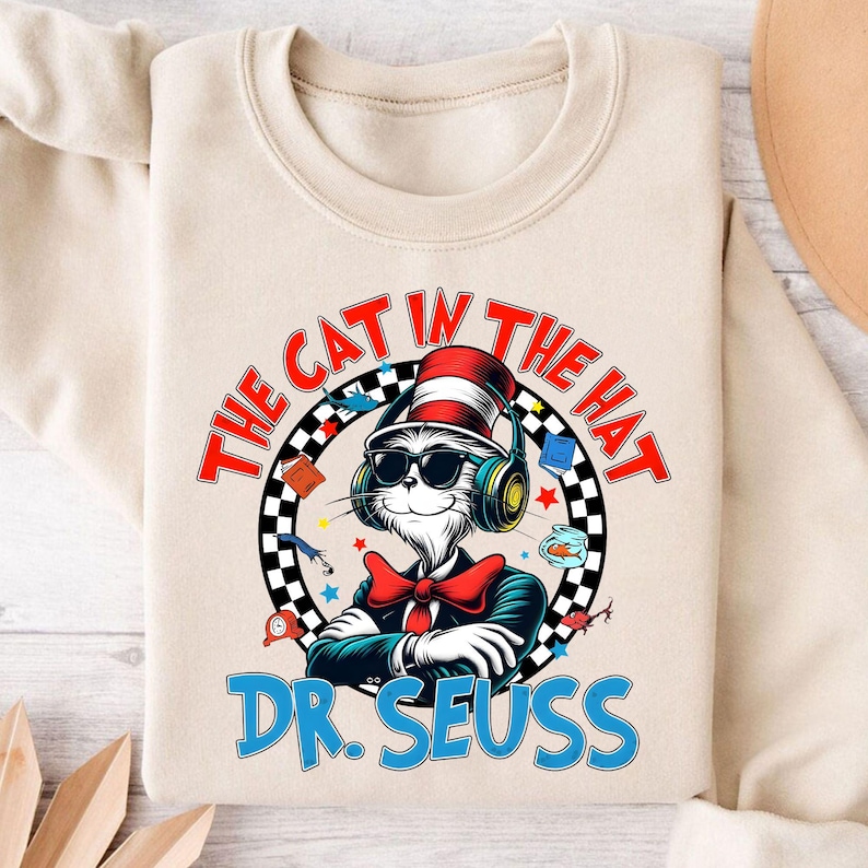 Peut inclure: Un sweat-shirt beige avec un imprim&eacute; graphique du Chat dans le Chapeau portant des lunettes de soleil et des &eacute;couteurs. Le texte "The Cat in the Hat" est dans un cercle rouge avec un motif &agrave; carreaux noir et blanc. Le texte "Dr. Seuss" est en bleu sous le cercle.