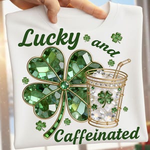 Puede incluir: Camiseta blanca con un diseño de trébol verde y las palabras "Lucky and Caffeinated". El trébol está hecho de gemas verdes, y también aparece una bebida con borde dorado con hielo y un trébol.