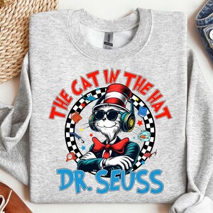 Peut inclure: Un sweat-shirt gris avec un motif repr&eacute;sentant le Chat dans le Chapeau portant des lunettes de soleil et des &eacute;couteurs. Le motif est entour&eacute; d'un cercle &agrave; damiers noir et blanc avec le texte "The Cat in the Hat" et "Dr. Seuss" en bleu.