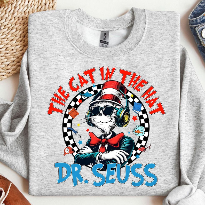 Peut inclure: Un sweat-shirt gris avec un motif repr&eacute;sentant le Chat dans le Chapeau portant des lunettes de soleil et des &eacute;couteurs. Le motif est entour&eacute; d'un cercle &agrave; damiers noir et blanc avec le texte "The Cat in the Hat" et "Dr. Seuss" en bleu.