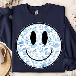 Könnte beinhalten: Ein marineblaues Sweatshirt mit einem weißen Smiley-Gesichtsdesign. Das Smiley-Gesicht ist mit einem blauen Aquarell-Blumenmuster gefüllt.