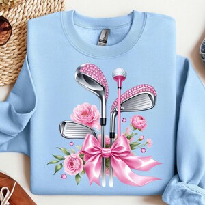 May include: Light blue sweatshirt with a pink and silver golf club design. The clubs are adorned with rhinestones and tied together with a pink bow. Pink flowers are arranged around the clubs.