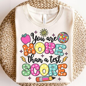 Peut inclure: T-shirt crème avec le texte "You are MORE than a test SCORE" en lettres à bulles colorées. Le motif comprend une pomme, un soleil, un globe, un crayon et des étoiles. Le t-shirt est en matière souple.