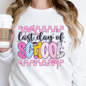 Puede incluir: Sudadera blanca con un gr&aacute;fico rosa, azul, amarillo y negro que dice "&uacute;ltimo d&iacute;a de escuela" con un lazo rosa y un patr&oacute;n a cuadros.