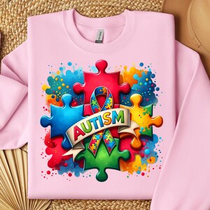 May include: Pink sweatshirt with a colorful puzzle piece design and the word "AUTISM" in a banner. The puzzle pieces are surrounded by colorful watercolor splatters.