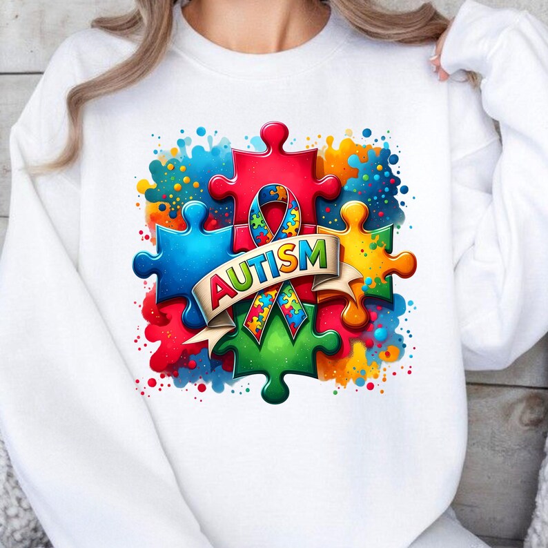 May include: White sweatshirt with a colorful puzzle piece design and the word "AUTISM" in a banner. The puzzle pieces are in shades of blue, red, green, yellow, and orange. The background is a white watercolor splatter.