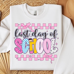 Puede incluir: Sudadera blanca con un gr&aacute;fico rosa, amarillo, azul y negro que dice "&uacute;ltimo d&iacute;a de escuela". El gr&aacute;fico est&aacute; decorado con un lazo, lunares y un patr&oacute;n a cuadros.