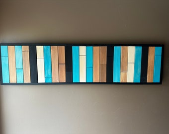 May include: A rectangular wooden wall art piece with a black frame. The art features vertical wooden planks in shades of blue, tan, and cream. The planks are arranged in a repeating pattern, creating a modern, geometric design.