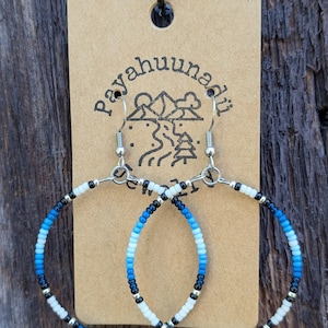May include: A pair of blue, white and black beaded hoop earrings with silver hooks. The earrings are displayed on a brown card with the text "Payahuunad" in black.