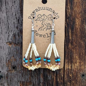 May include: A pair of dangle earrings with white, brown, and turquoise beads. The earrings are hanging from a brown card with the text "Payahuunadı Jewelry".