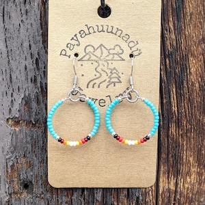 May include: A pair of turquoise beaded hoop earrings with a red, orange, yellow, white, and black bead design. The earrings are hanging on a brown card with the text "Payahuunad" printed on it.