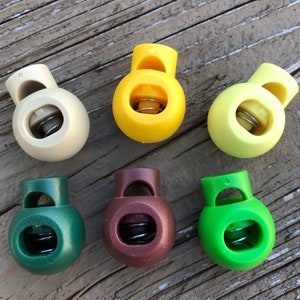 May include: Six round toggle cord locks in various colors, including yellow, beige, green, and brown. Each lock has a spring mechanism and a hole for threading cord.