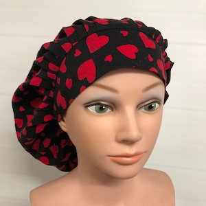 May include: A black scrub cap featuring a repeating pattern of distressed red hearts. The cap has a wide, flat front band and a gathered, pleated back section that provides a voluminous fit, covering the top and back of the head completely.