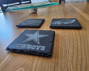 May include: Three black slate coasters with team logos. The coasters feature the logos for the Kansas City Chiefs, Philadelphia Eagles, and Dallas Cowboys. The coasters are on a wooden table.