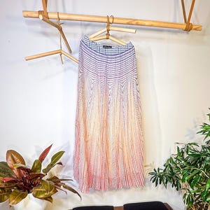 May include: A pleated skirt with a striped pattern in shades of red, orange, and white hangs on a wooden hanger. The skirt is suspended from a wooden rack. A bench with black cushions and plants in pots are in the foreground.