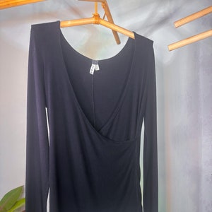 Puede incluir: Camiseta negra de manga larga con cuello en V y detalle de envoltura. La camiseta est&aacute; colgada en una percha de madera.