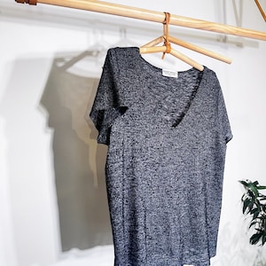 May include: A gray, short-sleeved, V-neck t-shirt hanging on a wooden rack with a leather strap. The shirt has a textured, slightly speckled fabric. The shirt has a small white tag with the text "FRONT ROW" on it.