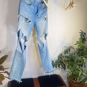 May include: Light blue denim jeans with distressed details, hanging from a wooden rod. The jeans have large rips on the front of the legs. The jeans are hanging above a wooden bench with two black cushions.