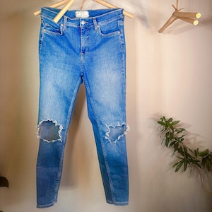 May include: Light wash blue denim jeans with distressed knees hanging on a wooden clothes hanger. The jeans are a skinny fit and have a mid-rise waist.  The brand label is visible on the back waistband.