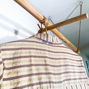 Op de afbeelding: Een gestreept shirt hangt aan een houten kledingrek. Het shirt is een lichtbruin en wit gestreept patroon.