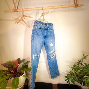 Op de afbeelding: Een lichtblauwe distressed denim jeans hangt aan een houten rek met leren banden. De jeans heeft verschillende scheuren en gaten in de stof.