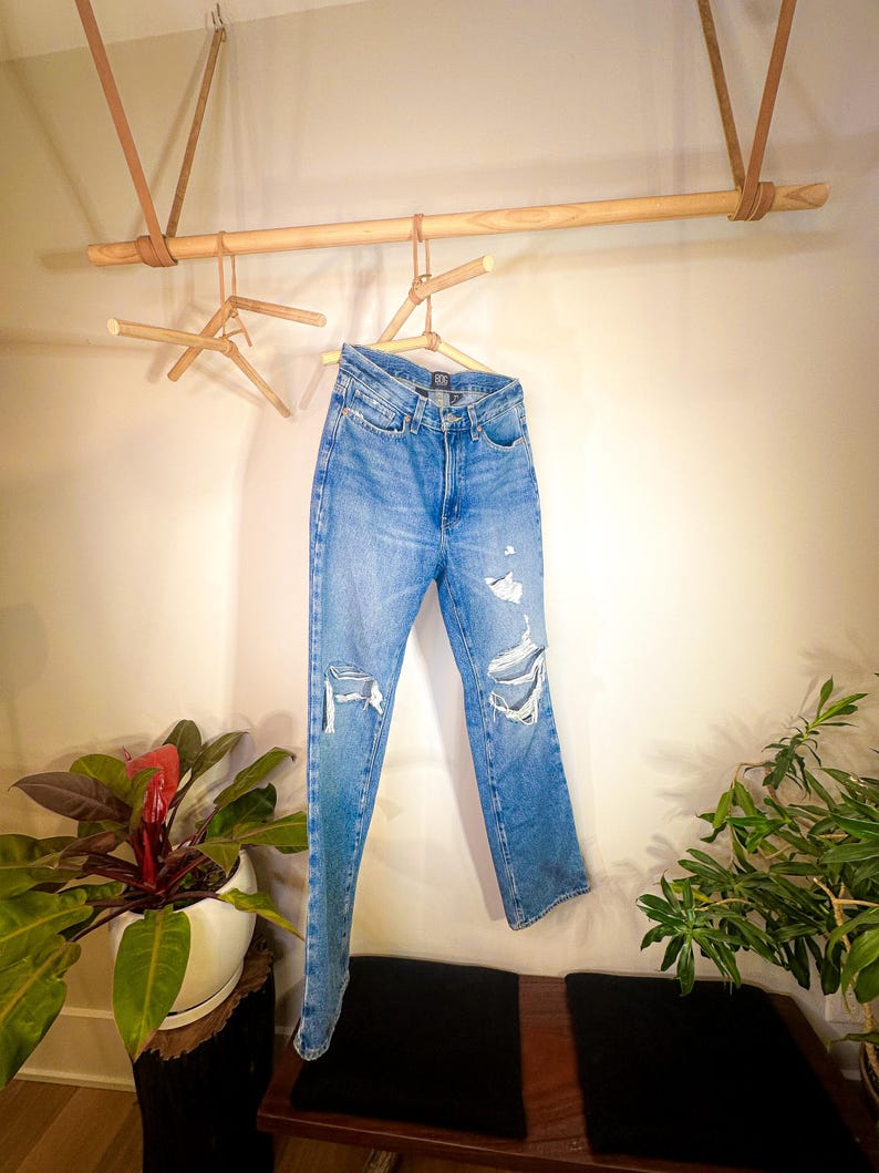 Op de afbeelding: Een lichtblauwe distressed denim jeans hangt aan een houten rek met leren banden. De jeans heeft verschillende scheuren en gaten in de stof.