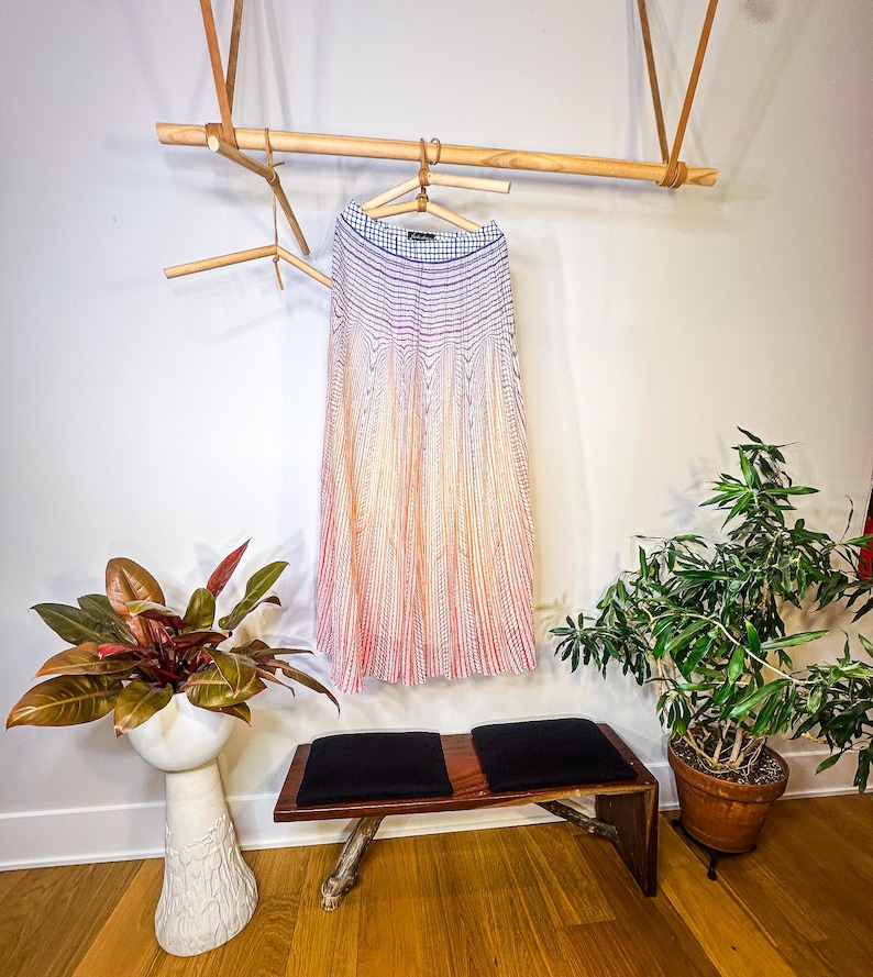 May include: A pleated skirt with a striped pattern in shades of red, orange, and white hangs on a wooden hanger. The skirt is suspended from a wooden rack. A bench with black cushions and plants in pots are in the foreground.