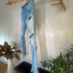 May include: Light blue denim jeans with distressed details, including ripped sections on the legs and frayed hems. The jeans are hanging on a wooden rod, showcasing their design and texture. The jeans are a light wash.