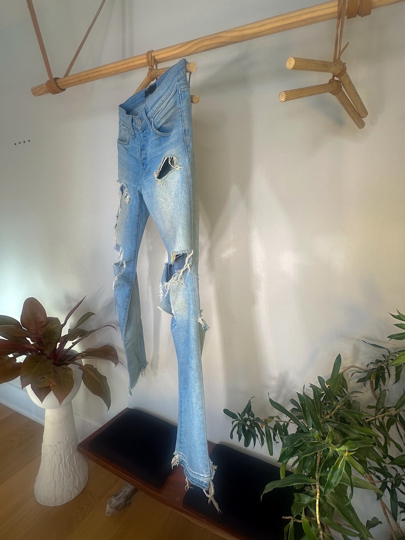 May include: Light blue denim jeans with distressed details, including ripped sections on the legs and frayed hems. The jeans are hanging on a wooden rod, showcasing their design and texture. The jeans are a light wash.