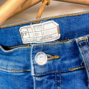 Op de afbeelding: Close-up van een blauwe spijkerbroek met een knoop en een label. Op het label staat "FP" en "MAAT: 23". De jeans hangt aan een houten hanger. Op de knoop staan de woorden "Free People".