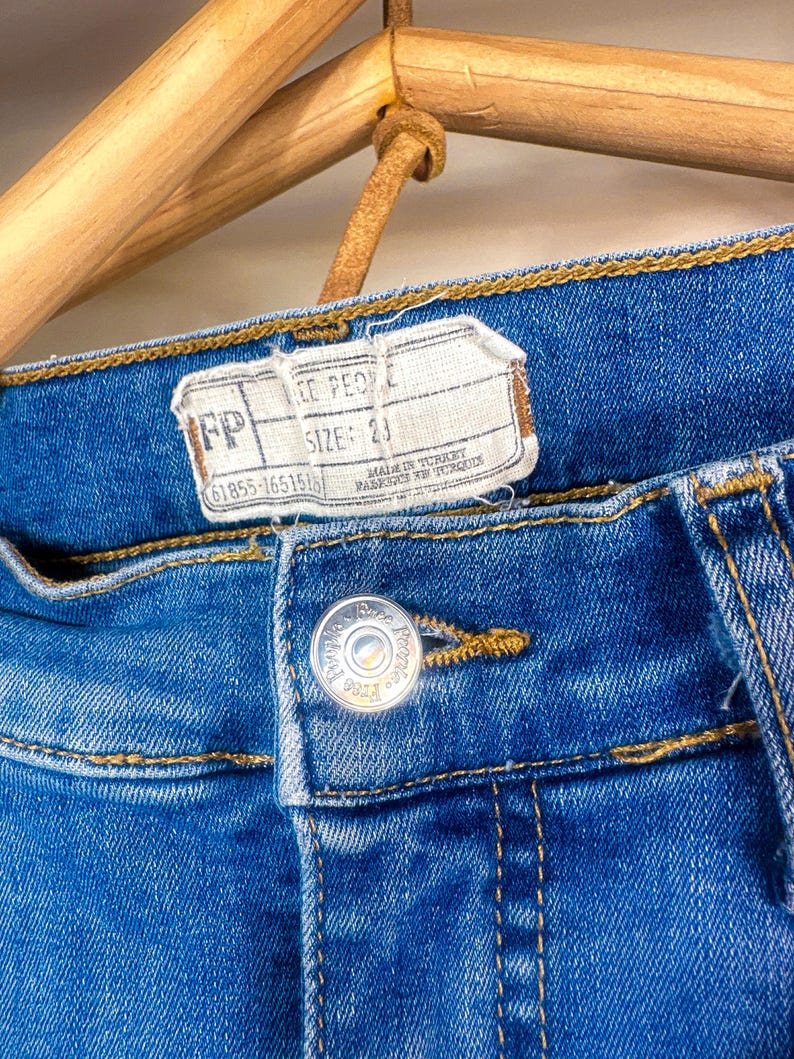 Op de afbeelding: Close-up van een blauwe spijkerbroek met een knoop en een label. Op het label staat "FP" en "MAAT: 23". De jeans hangt aan een houten hanger. Op de knoop staan de woorden "Free People".