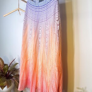May include: A pleated skirt with a gradient design, transitioning from blue at the top to orange and red at the bottom. The skirt is hanging on a wooden hanger, showcasing its vertical striped pattern.