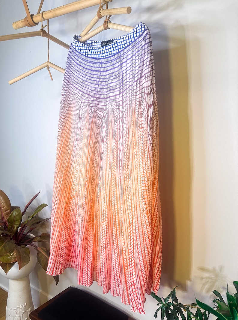 May include: A pleated skirt with a gradient design, transitioning from blue at the top to orange and red at the bottom. The skirt is hanging on a wooden hanger, showcasing its vertical striped pattern.